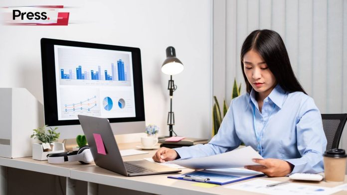 beautiful Asian woman working in office theme of white wearing blue shirt holding paper stack in hand and laptop connect with monitor showing bar charts pir charts and line charts with pink sticky note sticked on laptop