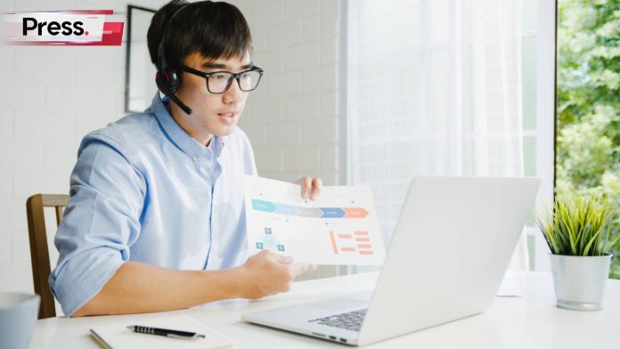 A young, bespectacled businessman is seated in his office, on a work video call with someone else. He is showing them a piece of paper with statistics and graphs on it, discussing their business performance and backlink building strategies.
