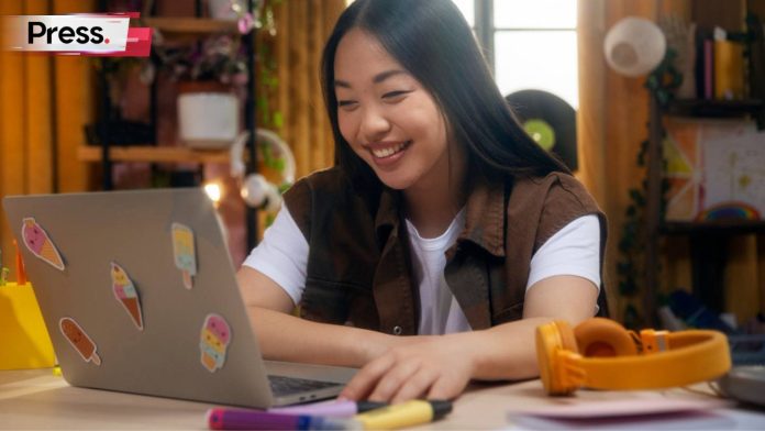 A female student sits at home, studying. She smiles as she looks at her laptop, which is covered in stickers. She is studying the role of AI in Malaysia.