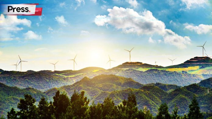 An image of a vista of rolling hills in the background, looming above a forest. On the tops of those hills, there are windmills that generate electricity, showcasing the importance of sustainability.