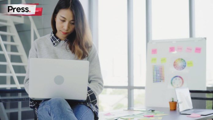 A serious-looking woman is typing on her laptop in a well-lit room, with a whiteboard behind her displaying all sorts of graphs and charts. She is analyzing her company's SEO and evaluating the keyword difficulty of various keywords on their website.