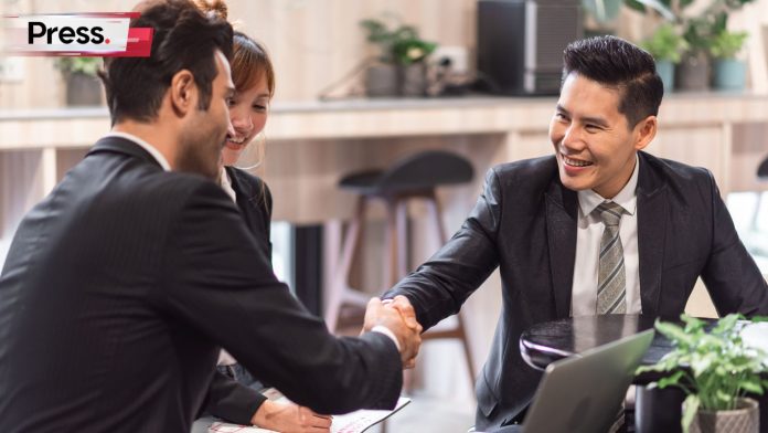 A group of two businessman and a businesswoman. All of them are smiling and happy. The businessmen are shaking hands and happily closing some sort of deal, possibly related to local SEO, with a laptop open on the table near them.