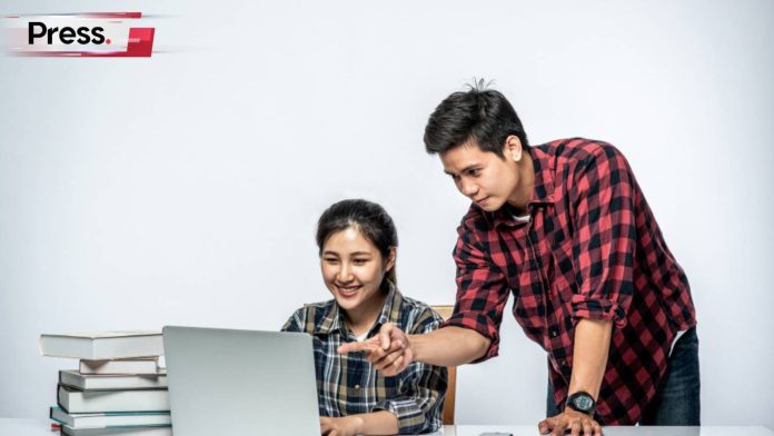 Two students using remote learning, one male one female, with the female student seated in front of a laptop. Next to the laptop is a pile of books, possibly belonging to her. She is smiling as the male student points at something on the laptop screen, instructing her on it.