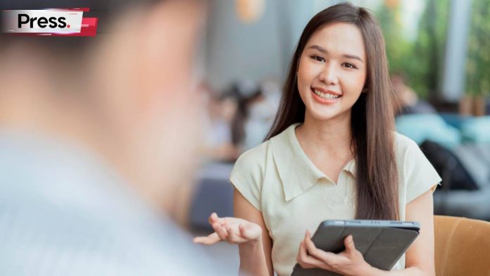 A pretty office lady in formal attire is standing in front of a man whose face we can't see. She is smiling at him while holding a laptop in one hand, and holding out her other hand as though suggesting something, reflecting that she is a good example of customer behavior.