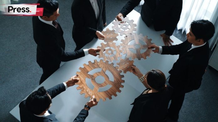 A group of professionals in formal attire collaborating at a table, connecting wooden gear pieces to symbolize coordinated teamwork and business alignment.