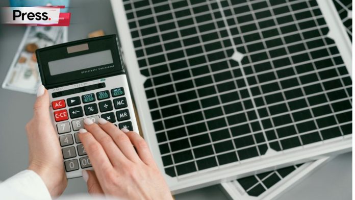 A photo of a person's hand holding a calculator, as the person stands in front of two solar panels which have been stacked atop each other on the ground. They are calculating the cost savings from this solar panel.