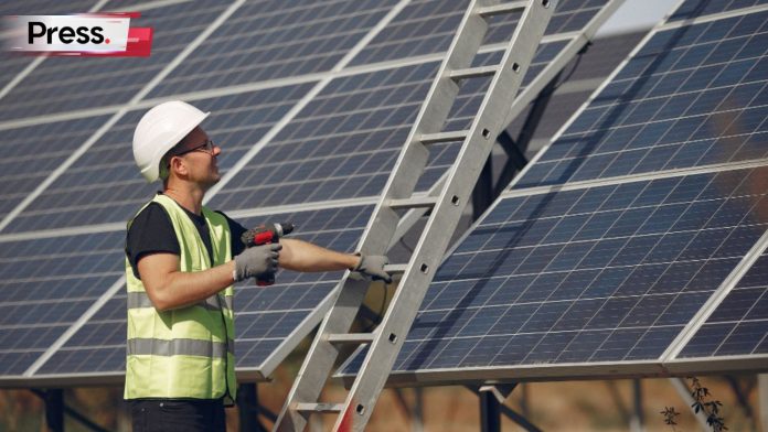 A construction worker performs a solar panel installation, holding on to his ladder as he does.