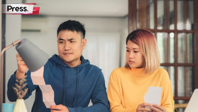A couple sits at a desk in their home, dressed comfortably in hoodies and casual wear. The husband is holding a paper in his hands, his PTPTN loan, and his wife looks on with a concerned frown.