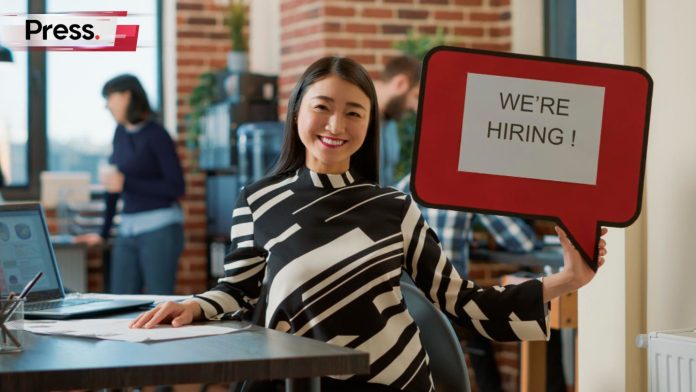 An office worker seated at her desk, holding up a sign saying 'We're Hiring', which represents the debate between hiring in-house or an external SEO agency in Malaysia.