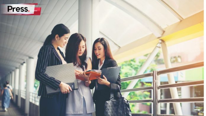 Three girls, who may be tertiary education students in Malaysia, are dressed neatly and stand close by each other, discussing some documents.